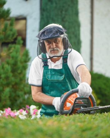 Älterer Mann in Schutzhelm und grüner Latzhose bedient eine Motorsäge im Garten
