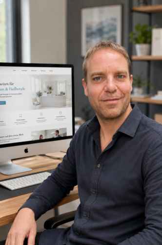 Mann sitzt am Schreibtisch vor einem Computer und schaut in die Kamera