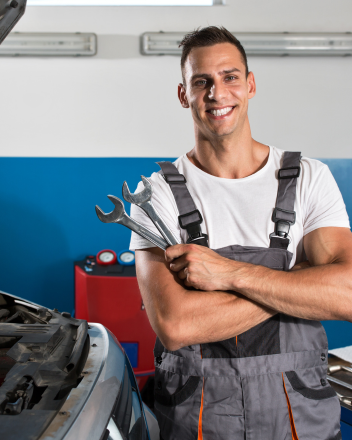 Lächelnder Mechaniker mit Schraubenschlüsseln vor einer geöffneten Motorhaube in der Werkstatt