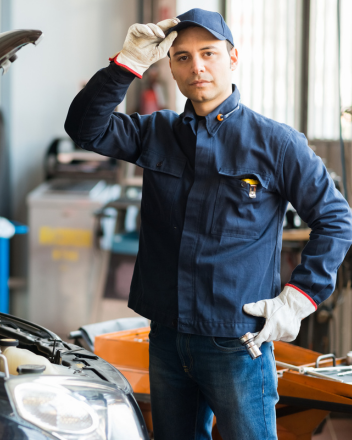 Mechaniker in blauer Arbeitskleidung und Handschuhen steht neben einem Auto und hebt die Mütze an