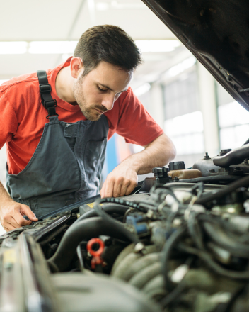 Mechaniker in rotem Shirt und Latzhose prüft den Motorraum eines Autos unter geöffneter Motorhaube