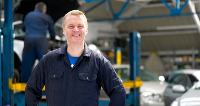 Mann in Werkstattkleidung lächelt vor einer Autowerkstatt, im Hintergrund arbeitet ein Kollege an einem Hebebühnenwagen