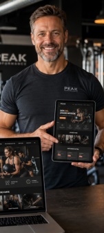 Lächelnder Mann im Gym hält ein Tablet mit Fitness-App und zeigt auf den Bildschirm; Laptop daneben zeigt dieselbe App